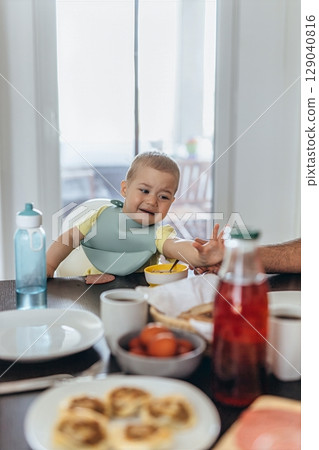 Toddler reaching for food on breakfast table. Child sits in a high chair with family enjoying a cozy morning meal at home. Bright kitchen scene with natural light 129040816