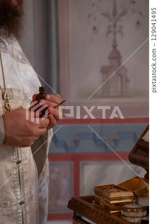 Orthodox priest prepares holy oil for the anointing ritual during a baptism ceremony, holding a vial and brush Orthodox priest prepares holy oil for the anointing ritual during a baptism ceremony, holding a vial and brush 129041495