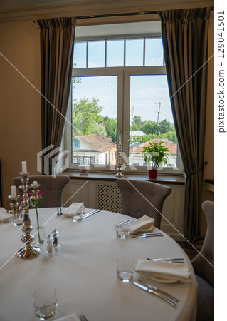 Elegant restaurant interior with a round table set for six near a large window overlooking old town rooftops and trees 129041501