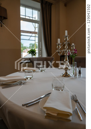 Close-up of a beautifully set round table in an elegant restaurant, featuring white linen, silver cutlery, glassware, and a silver candelabra Close-up of a beautifully set round table in an elegant restaurant, featuring white linen, silver cutlery, glassware, and a silver candelabra 129041502