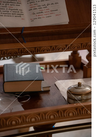 Close-up of an Orthodox prayer book, placed on a carved wooden lectern inside a church, accompanied by a small ornate container and baptismal crosses 129041513