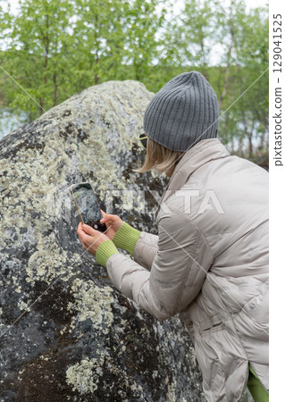 A woman photographs a moss-covered boulder in a lush northern forest using her smartphone 129041525