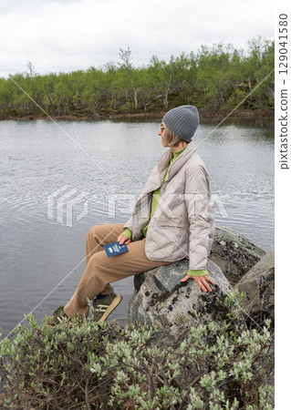 A woman in warm clothes and sunglasses sits on a rock by a cold northern lake, holding a polar explorer passport in her hand 129041580