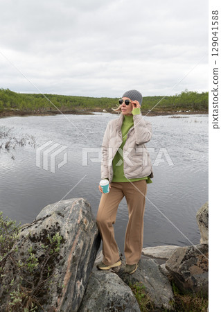 Woman in a beige puffer jacket and gray beanie stands by an Arctic lake holding a turquoise takeaway coffee cup Woman in a beige puffer jacket and gray beanie stands by an Arctic lake holding a turquoise takeaway coffee cup 129041588