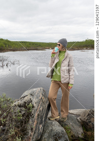 Woman in beige jacket and gray beanie drinks from a turquoise coffee cup while standing on rocks by a northern lake in Teriberka, Russia 129041597