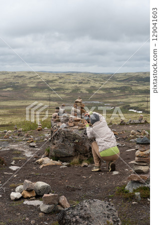 Woman in a grey beanie and light jacket photographs stacked stone cairns on a mossy tundra plateau near Teriberka, Russia 129041603