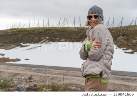 Cheerful woman in a grey beanie and sunglasses smiling and hugging herself to stay warm on a snowy roadside in Teriberka, Russia 129041609