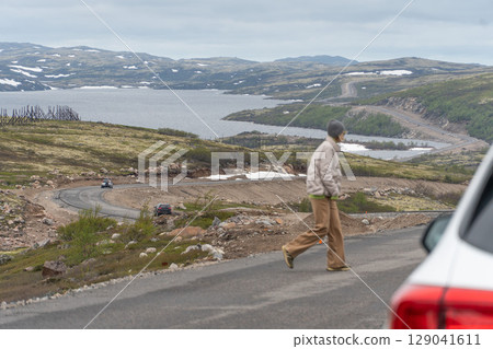 A woman in a gray beanie and beige jacket crosses a winding road surrounded by the Arctic tundra landscape near Teriberka, Russia A woman in a gray beanie and beige jacket crosses a winding road surrounded by the Arctic tundra landscape near Teriberka, Russia 129041611