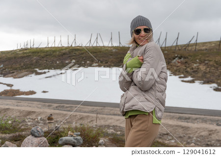 Cheerful woman in a grey beanie and sunglasses smiling and hugging herself to stay warm on a snowy roadside in Teriberka, Russia Cheerful woman in a grey beanie and sunglasses smiling and hugging herself to stay warm on a snowy roadside in Teriberka, Russia 129041612