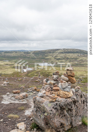Multiple stone cairns are carefully balanced on a rocky tundra plateau overlooking mossy hills, lakes, and snowy patches near Teriberka, Russia 129041618
