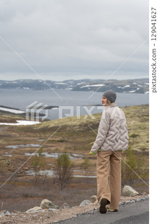 Woman in beige jacket and gray beanie walking along a road overlooking Arctic tundra and Barents Sea with snow patches on distant hills 129041627
