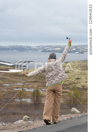 Joyful woman in beige jacket and gray beanie raising arms with sunglasses in hand, enjoying the dramatic Arctic scenery with snowy hills 129041632