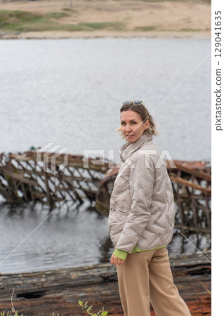 Woman in a beige puffer jacket and sunglasses standing near rusted shipwrecks by the shore, looking at the camera with a calm expression 129041635
