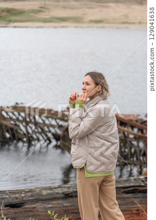 Woman in a beige puffer jacket and sunglasses standing near rusted shipwrecks by the shore, looking at the camera with a calm expression 129041638