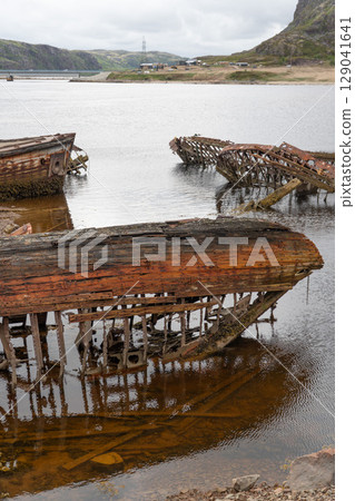 Rusted wooden and metal shipwrecks partially submerged in shallow coastal waters against a backdrop of rocky hills and cloudy skies Rusted wooden and metal shipwrecks partially submerged in shallow coastal waters against a backdrop of rocky hills and cloudy skies 129041641