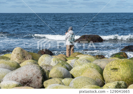 Woman in white jacket photographing the Barents Sea while standing on Dragon Eggs Beach, Teriberka, with large round green boulders in the foreground Woman in white jacket photographing the Barents Sea while standing on Dragon Eggs Beach, Teriberka, with large round green boulders in the foreground 129041665