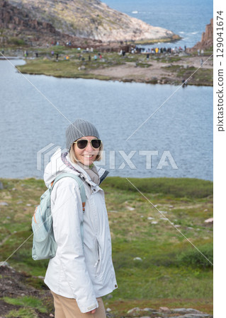 Smiling woman in a white jacket and gray beanie standing by a serene lake, with cliffs and the Barents Sea visible in the background 129041674