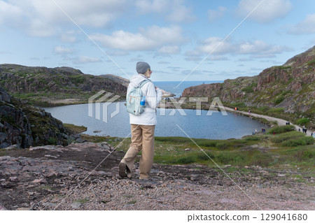 Woman with a backpack and water bottle looking at the scenic Arctic coastline and cliffs in Teriberka Woman with a backpack and water bottle looking at the scenic Arctic coastline and cliffs in Teriberka 129041680