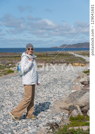 Woman in a white jacket and gray beanie holding a light blue backpack with a yellow rain cover, standing on a rocky path in Teriberka nature reserve Woman in a white jacket and gray beanie holding a light blue backpack with a yellow rain cover, standing on a rocky path in Teriberka nature reserve 129041681