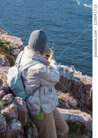 Woman in warm clothing sitting on a rocky Arctic cliff at the Barents Sea in Teriberka park, photographing a seabird colony with her smartphone 129041718