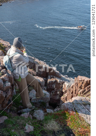 Woman with backpack and winter clothing standing on a rocky cliff by the Barents Sea in Teriberka, photographing a boat and seabird colony below 129041721