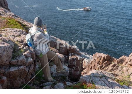 Woman with backpack and winter clothing standing on a rocky cliff by the Barents Sea in Teriberka, photographing a boat and seabird colony below 129041722