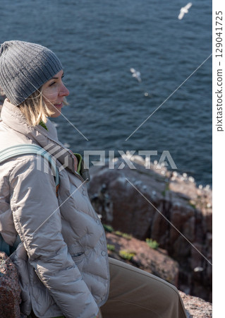 Smiling woman in a gray beanie and light down jacket standing on a rocky Arctic cliff above the Barents Sea, observing a seabird colony below 129041725