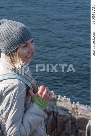 Smiling woman in a gray beanie and light down jacket standing on a rocky Arctic cliff above the Barents Sea, observing a seabird colony below Smiling woman in a gray beanie and light down jacket standing on a rocky Arctic cliff above the Barents Sea, observing a seabird colony below 129041726
