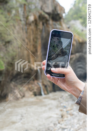 A womans hand holding a smartphone and taking a photo of the scenic Chegem Gorge with a waterfall 129041759