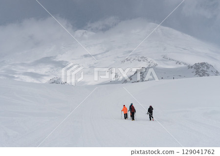 Group of climbers with trekking poles and backpacks ascending snow-covered Mount Elbrus under dramatic cloudy skies 129041762