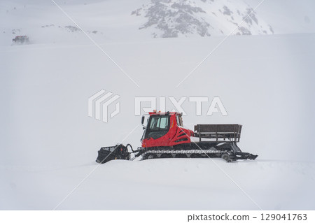 Red snow groomer operating on the snowy slopes of Mount Elbrus in heavy snowfall, surrounded by pristine white snow 129041763