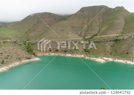A vivid turquoise mountain lake winds through the lush, mist-covered hills of the North Caucasus 129041792