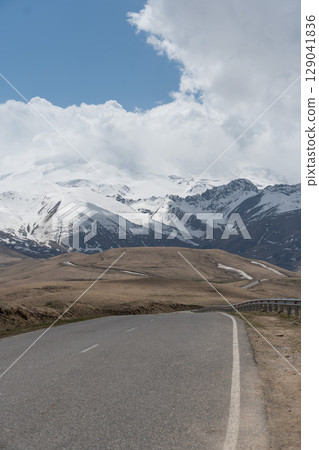 Empty winding mountain road leading toward snow-covered peaks of Mount Elbrus under dramatic skies. Editorial use: road trip, local tourism Empty winding mountain road leading toward snow-covered peaks of Mount Elbrus under dramatic skies. Editorial use: road trip, local tourism 129041836