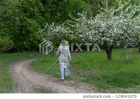 Woman in a plaid coat walks through a blooming orchard. Mindfulness, seasonal change, slow living, and mental well-being 129041870