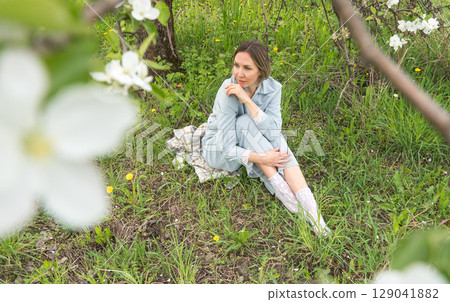 Middle-aged woman in soft blue loungewear sits on a checkered blanket in a blooming orchard. Mindfulness, spa retreats, slow fashion, mental health therapy, and natural beauty marketing Middle-aged woman in soft blue loungewear sits on a checkered blanket in a blooming orchard. Mindfulness, spa retreats, slow fashion, mental health therapy, and natural beauty marketing 129041882