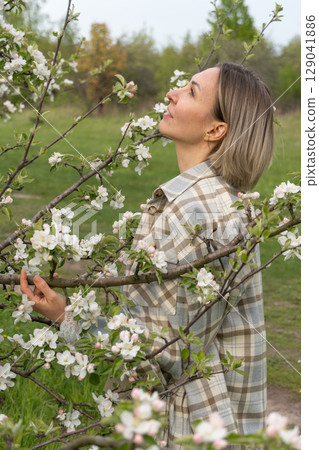 Woman in a plaid coat standing among blooming branches in a spring orchard, softly gazing through apple blossoms. Femininity, self-connection, beauty in nature, and spring renewal Woman in a plaid coat standing among blooming branches in a spring orchard, softly gazing through apple blossoms. Femininity, self-connection, beauty in nature, and spring renewal 129041886