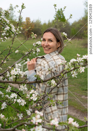 Woman in a plaid coat standing among blooming branches in a spring orchard, softly gazing through apple blossoms. Femininity, self-connection, beauty in nature, and spring renewal 129041889