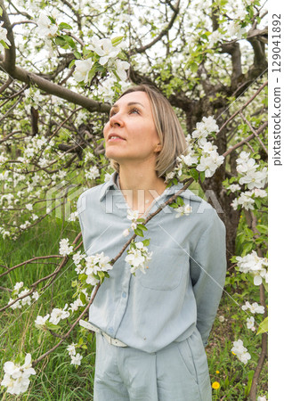 A woman in a light blue outfit stands among blooming apple trees. Mindfulness, connection with nature, mental well-being, and eco-lifestyle A woman in a light blue outfit stands among blooming apple trees. Mindfulness, connection with nature, mental well-being, and eco-lifestyle 129041892