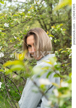 Smiling middle-aged woman in a light blue outfit standing among blooming branches. Natural cosmetics, age positivity, emotional wellness Smiling middle-aged woman in a light blue outfit standing among blooming branches. Natural cosmetics, age positivity, emotional wellness 129041905