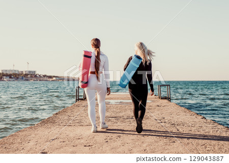 Yoga Women Beach Pier - Two women walk on a pier toward the ocean carrying yoga mats, enjoying a sunrise or sunset. Yoga Women Beach Pier - Two women walk on a pier toward the ocean carrying yoga mats, enjoying a sunrise or sunset. 129043887
