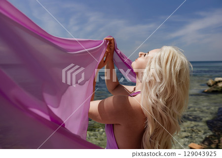 Woman Beach Fashion: Blonde woman holds purple fabric to sky at beach for sunny vacation shoot. 129043925