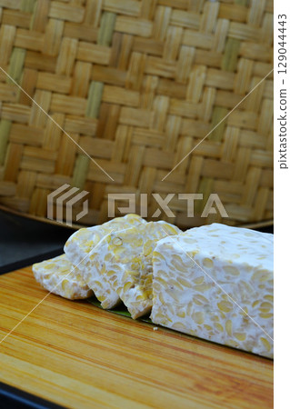 A block of tempeh, partially sliced, sits on a wooden cutting board with a woven bamboo mat in the background. 129044443
