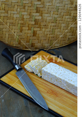 A block of tempeh, partially sliced, on a wooden cutting board with a knife, set against a woven bamboo background. 129044826