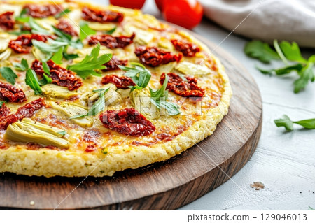 A delicious cauliflower crust pizza is topped with sun-dried tomatoes, artichokes, and vegan parmesan. The cozy kitchen setting features rustic wooden countertops lit by warm morning sunlight 129046013