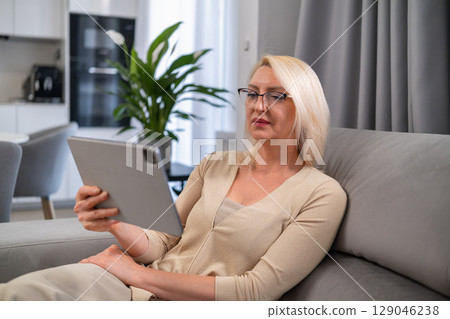 Smiling middle aged lady enjoying a film on her tablet, seated on a cozy sofa at home. Entertainment, leisure and modern technology concept. Smiling middle aged lady enjoying a film on her tablet, seated on a cozy sofa at home. Entertainment, leisure and modern technology concept. 129046238
