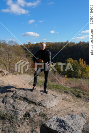 Beautiful blonde man playing acoustic guitar and singing relaxing enjoying outdoors on sunny autumn day. Artist learning and practicing playing musical instrument. Fall leaves autumnal 129046462
