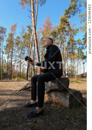 Portrait of smiling caucasian artist plays wooden acoustic guitar in autumnal park. Young songwriter plays string musical instrument hobby outside in nature fall time. Audio music healing Portrait of smiling caucasian artist plays wooden acoustic guitar in autumnal park. Young songwriter plays string musical instrument hobby outside in nature fall time. Audio music healing 129046463