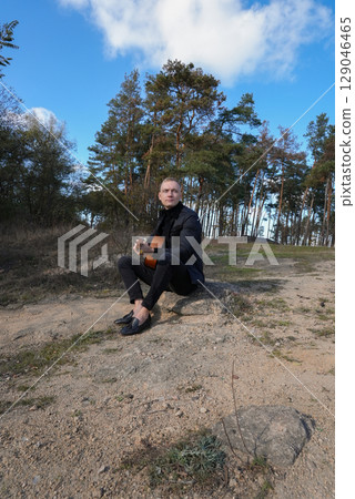 Portrait of smiling caucasian artist plays wooden acoustic guitar in autumnal park. Young songwriter plays string musical instrument hobby outside in nature fall time. Audio music healing 129046465
