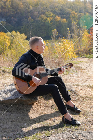 Blonde man playing string guitar outdoors in autumn forest. Concept of sound therapy, mental health and wellness rituals. Calmness tranquility audio-sensory practices. Aura farming energy 129046467