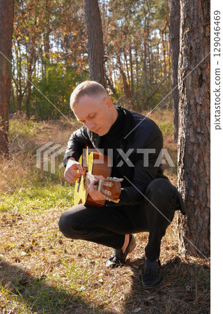 Blonde man playing string guitar outdoors in autumn forest. Concept of sound therapy, mental health and wellness rituals. Calmness tranquility audio-sensory practices. Aura farming energy 129046469